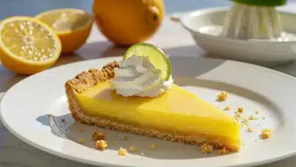 Part de tarte au citron brillante avec chantilly et zeste de citron vert sur assiette blanche, sous la lumière du soleil.