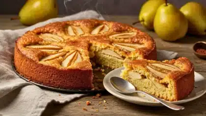 Gâteau moelleux aux poires doré, avec des lamelles de poires caramélisées sur le dessus, présenté sur une table en bois, une part servie dans une assiette avec une cuillère.